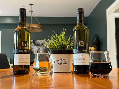 Two Gray Monk wine bottles and glasses flank a potted plant on a wooden table.