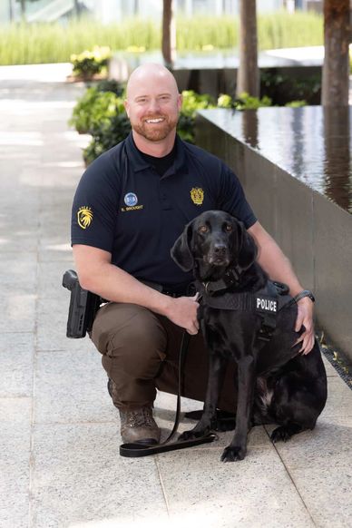 Police officer kneels next to a black police dog by a water feature.