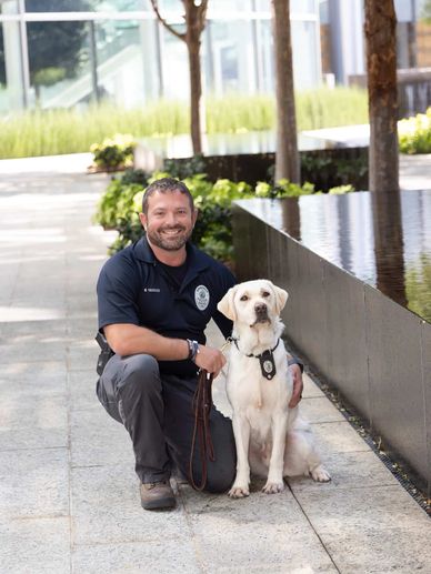 Police officer kneels beside his K-9 unit dog outdoors near a reflective water feature.