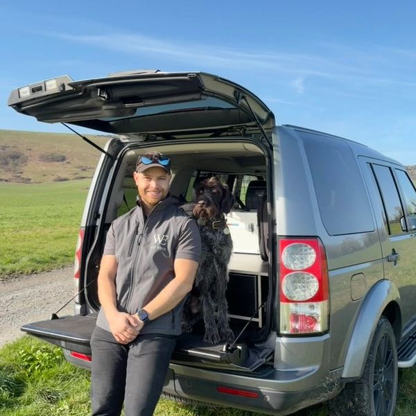 Man and dog sitting in the open trunk of an SUV in a grassy area.