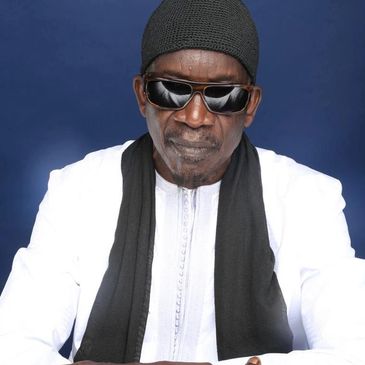 Man in white traditional attire, black scarf, and sunglasses.
