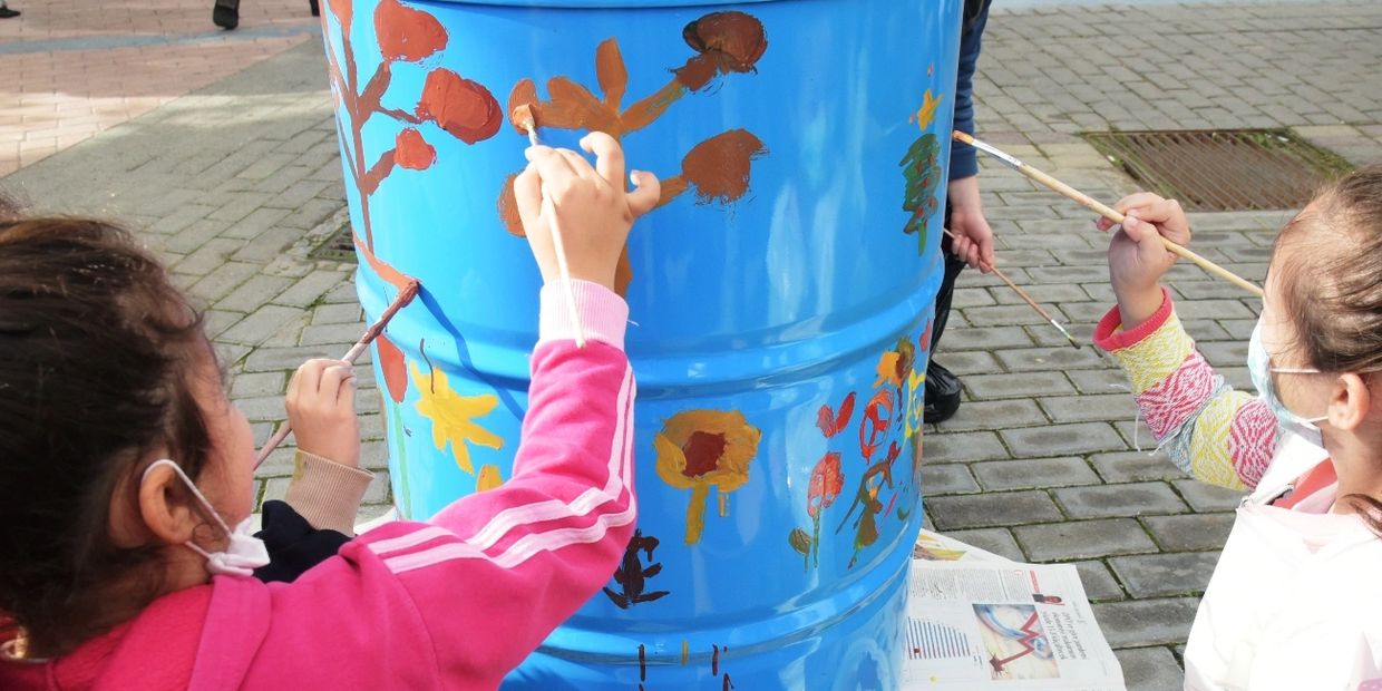 Children painting colorful designs on a blue barrel outdoors.