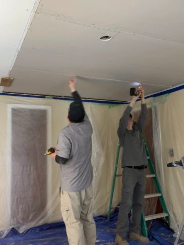 Two men installing drywall on a ceiling in a room under renovation.