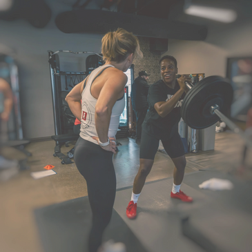 Two people working out together in a gym, one lifting weights and the other watching.