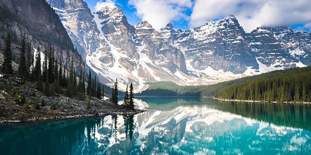 Snow-capped mountains reflecting on a clear blue lake surrounded by pine trees.