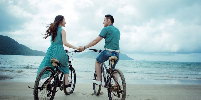 Couple holding hands while riding bicycles on the beach.