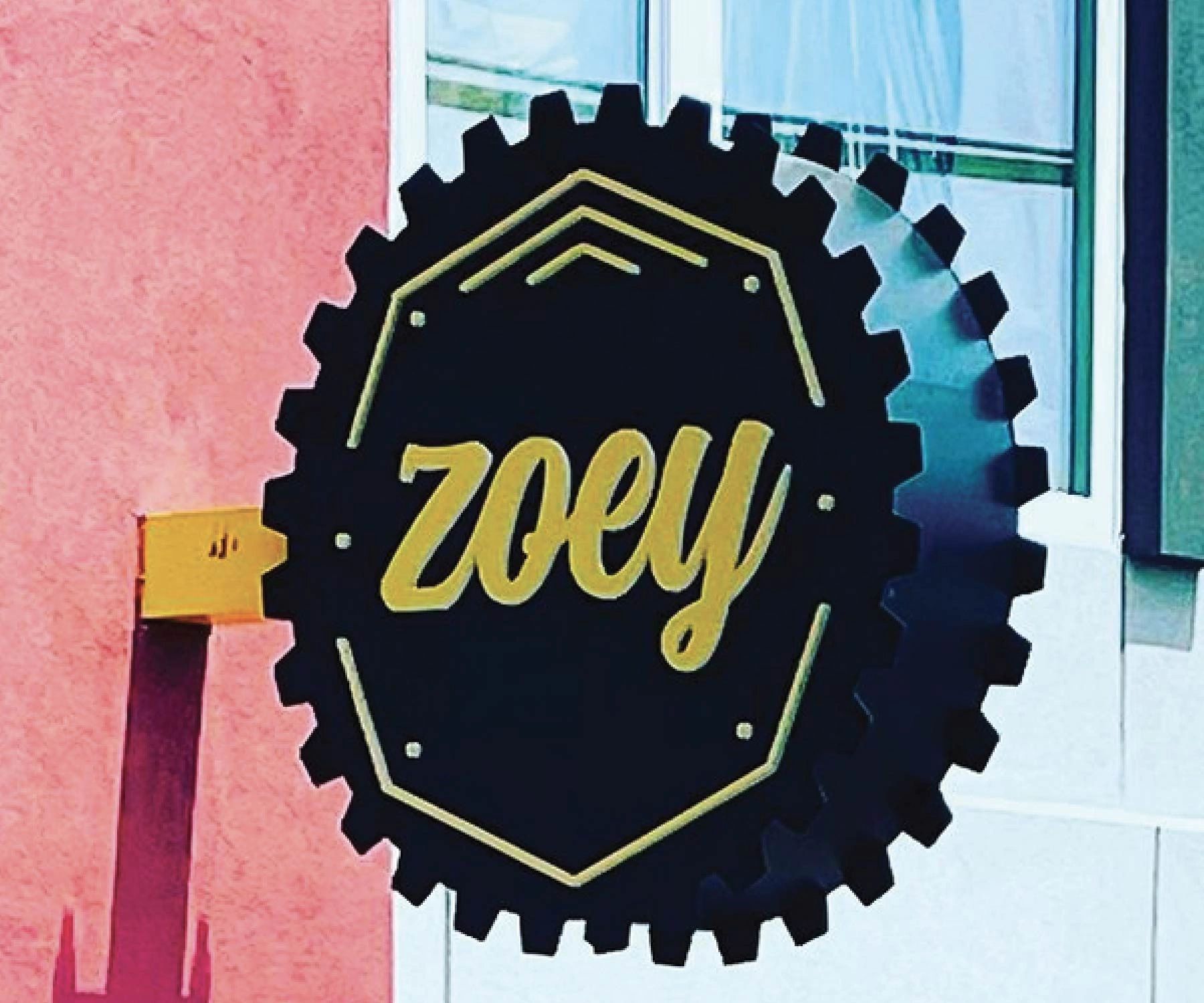 Black gear-shaped sign with 'zoey' in gold letters on a building.