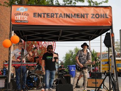Live band performing under Grilled Cheese Challenge's Entertainment Zone tent outdoors.