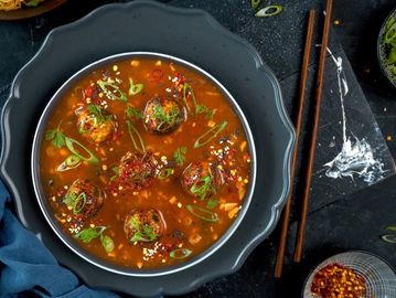 Bowl of spicy soup with meatballs and garnished with herbs and chili flakes.