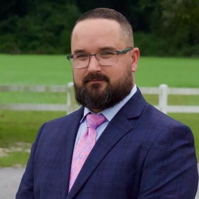 Man in a navy suit and pink tie standing outdoors with trees and a white fence in the background.