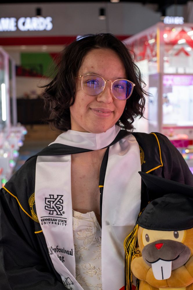 a picture of tiffany in an arcade wearing a black masters gown. and holding a brown plushie