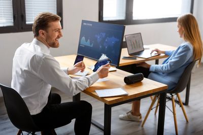 Two colleagues working at desks with laptops and charts in an office.