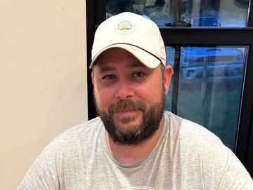 Smiling man with a beard and cap sitting indoors near a window.