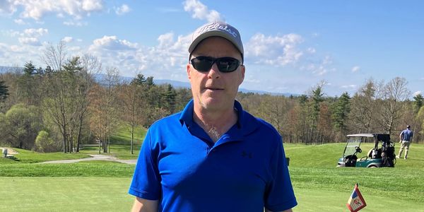 Man in blue polo and cap standing on a golf course on a sunny day.