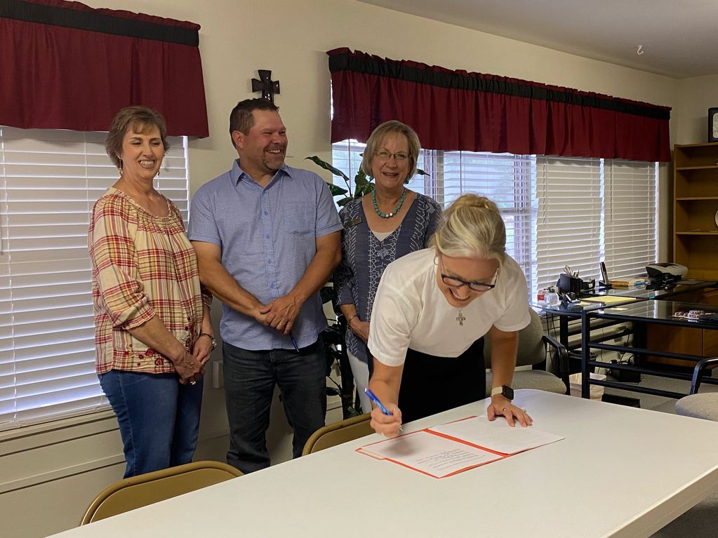 A woman signs a document while three others watch and smile in a well-lit room.