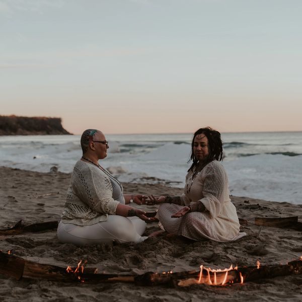 Two people meditate inside a circle of burning wood on the beach at sunset.