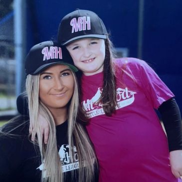 Two smiling girls wearing matching MH baseball caps and sports shirts.