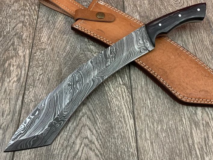 A handmade knife with a 15-inch overall length and a 10-inch blade made of Damascus steel, composed of 15n20 and 1095. The handle is made of densified wood and the knife is accompanied by a hand-stitched cowhide leather sheath with a belt loop. The knife weighs approximately 434 grams and has a Rockwell hardness rating of 55-58. 