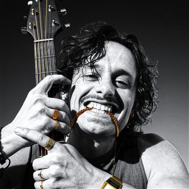 Man smiling with guitar and gold jewelry in black and white photo.