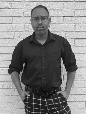 Man in glasses and black shirt standing against a white brick wall.