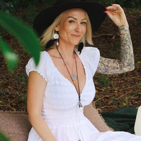 A woman in a white dress and black hat smiles outdoors among greenery.