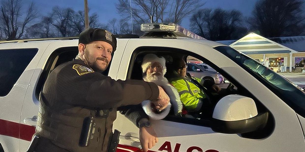Santa Claus sitting in a police car with officers at dusk.
