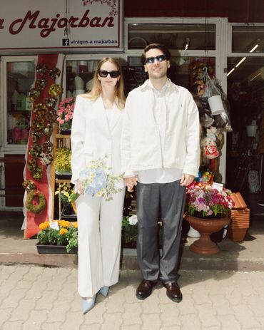 Couple in white outfits holding hands outside a flower shop with bouquet.