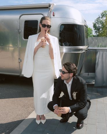 Stylish couple in formal attire enjoying drinks outside a shiny trailer on a sunny day.