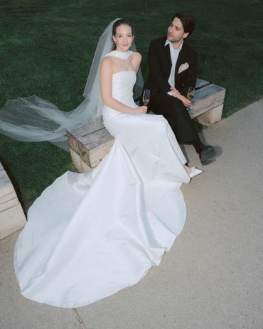 Bride in white gown and groom in black suit sitting on a bench with champagne glasses.