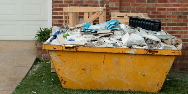 A yellow dumpster filled with construction debris sits outside a brick house.