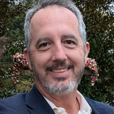 Smiling man with salt-and-pepper beard outdoors near flowers.