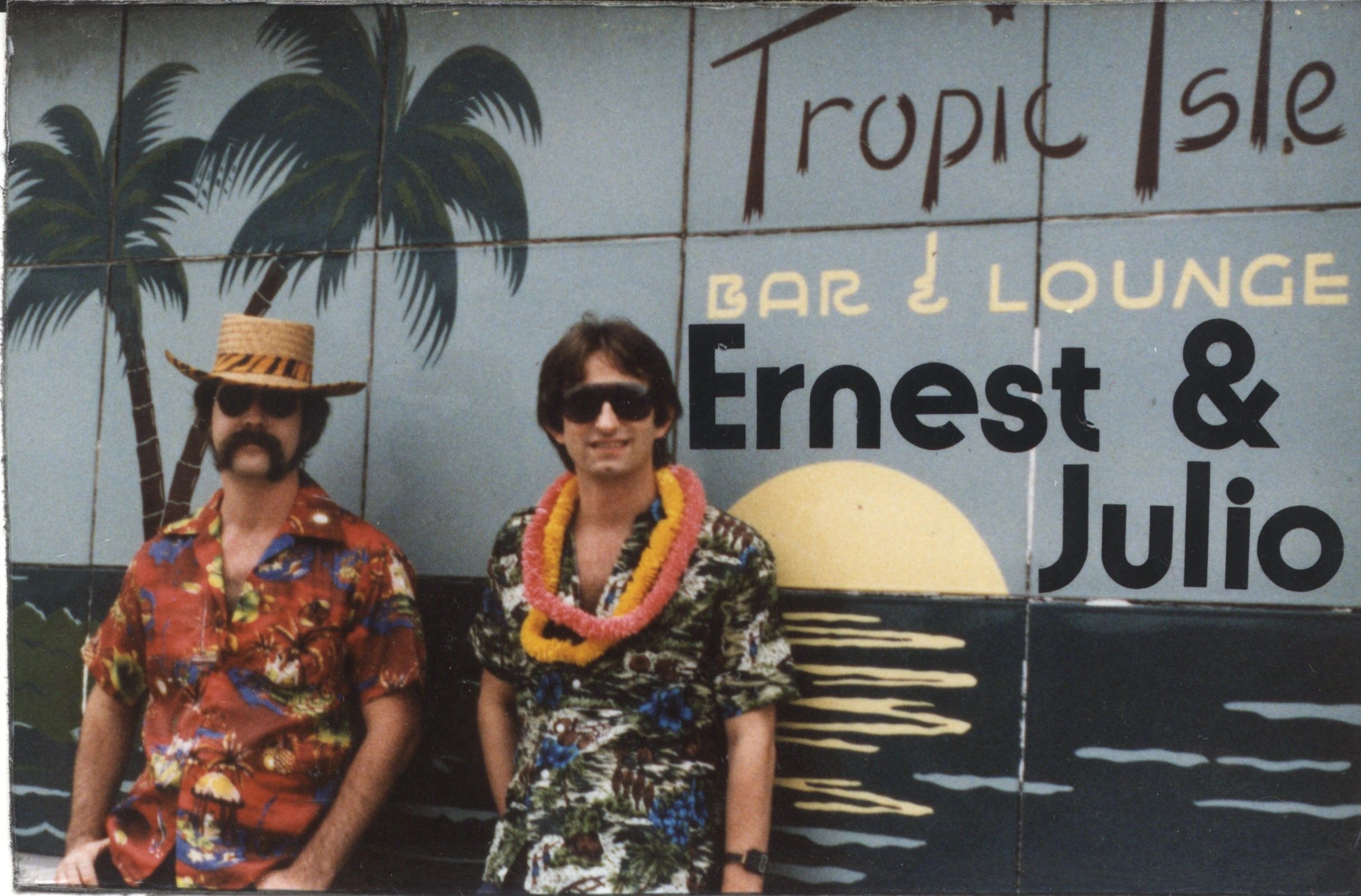 Two men in tropical shirts and sunglasses in front of a bar mural.