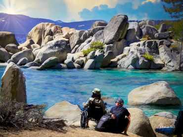 A woman in a wheelchair beside a man squatting beside her on the beach in Lake Tahoe.