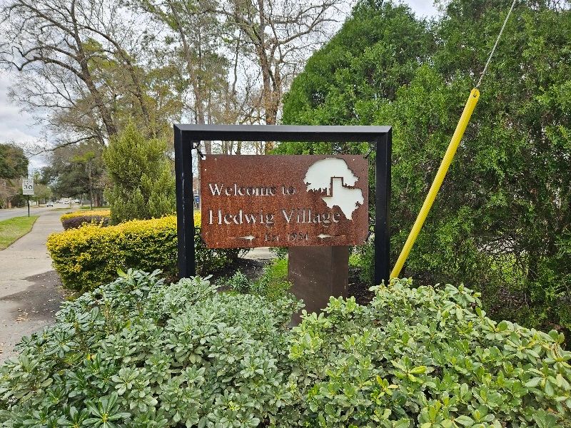 Welcome to Hedwig Village sign with rust AND a black frame. Green and yellow trees and shrubs.