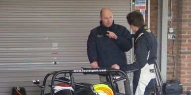Two men in racing suits having a discussion near a garage door.