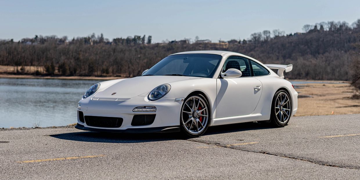 White Porsche sports car parked by a lake with a forest backdrop.