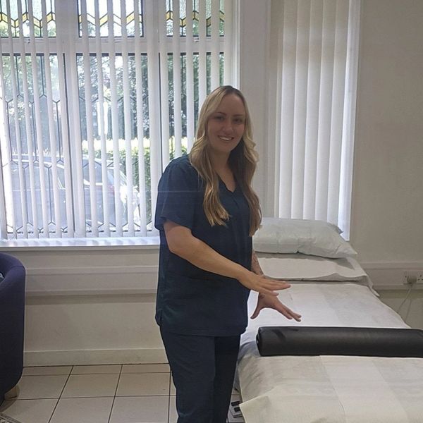A smiling woman in medical scrubs stands beside a treatment bed in a bright room.