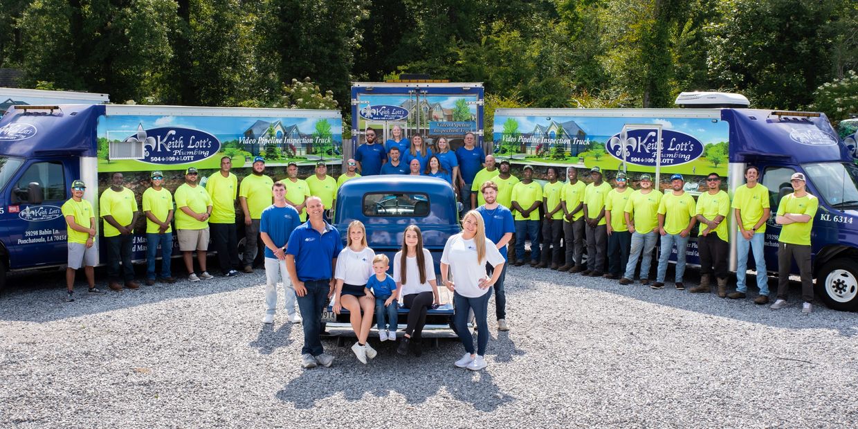 Large plumbing company team posing with branded trucks and a vintage blue car outdoors.