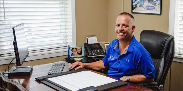 Man in blue polo shirt sitting at a wooden office desk with a computer and calendar.