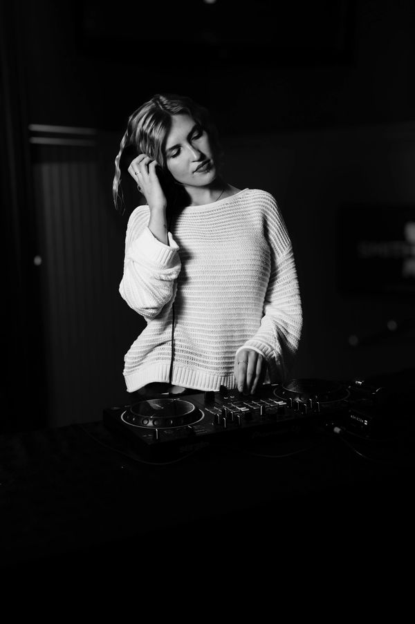 Woman DJ wearing headphones, adjusting music on a mixer in a dimly lit room.