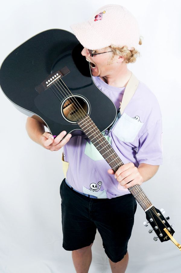Person wearing a hat playfully biting a black acoustic guitar.