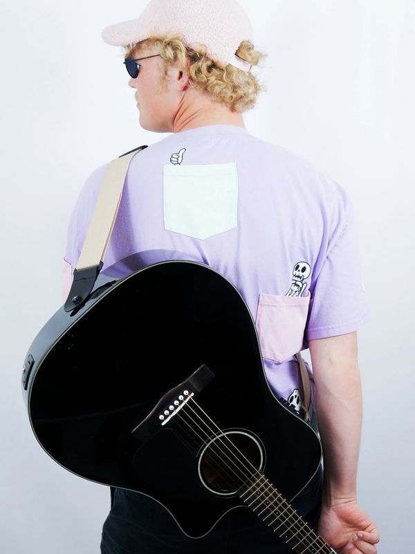 Person with curly hair wearing a pastel shirt and carrying a black acoustic guitar.