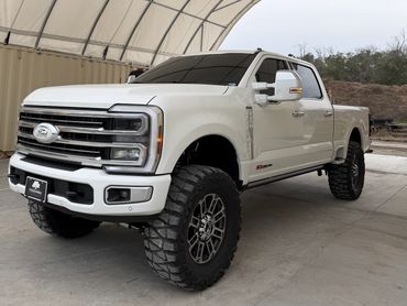 White lifted Ford pickup truck with large off-road tires under a canopy.