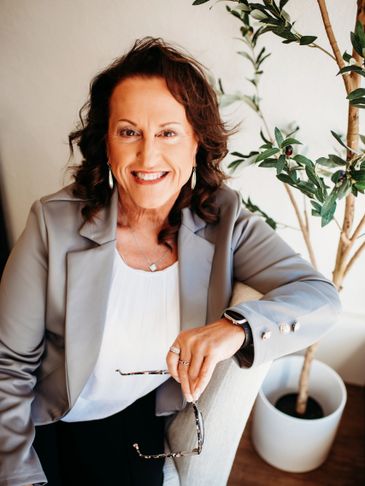 Smiling woman in blazer holding glasses, sitting by a plant.