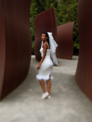 Bride in white dress and veil walking between large curved sculptures outdoors.