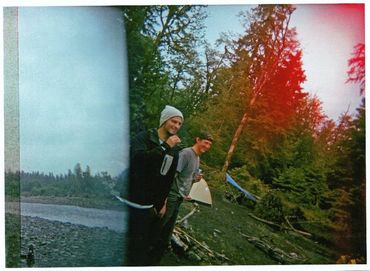 Two young men standing and smiling by a wooded riverbank with a surfboard nearby.