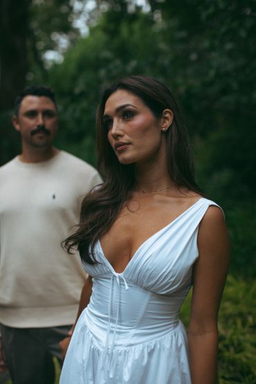 Woman in a white dress with a man blurred in the background in a forest setting.