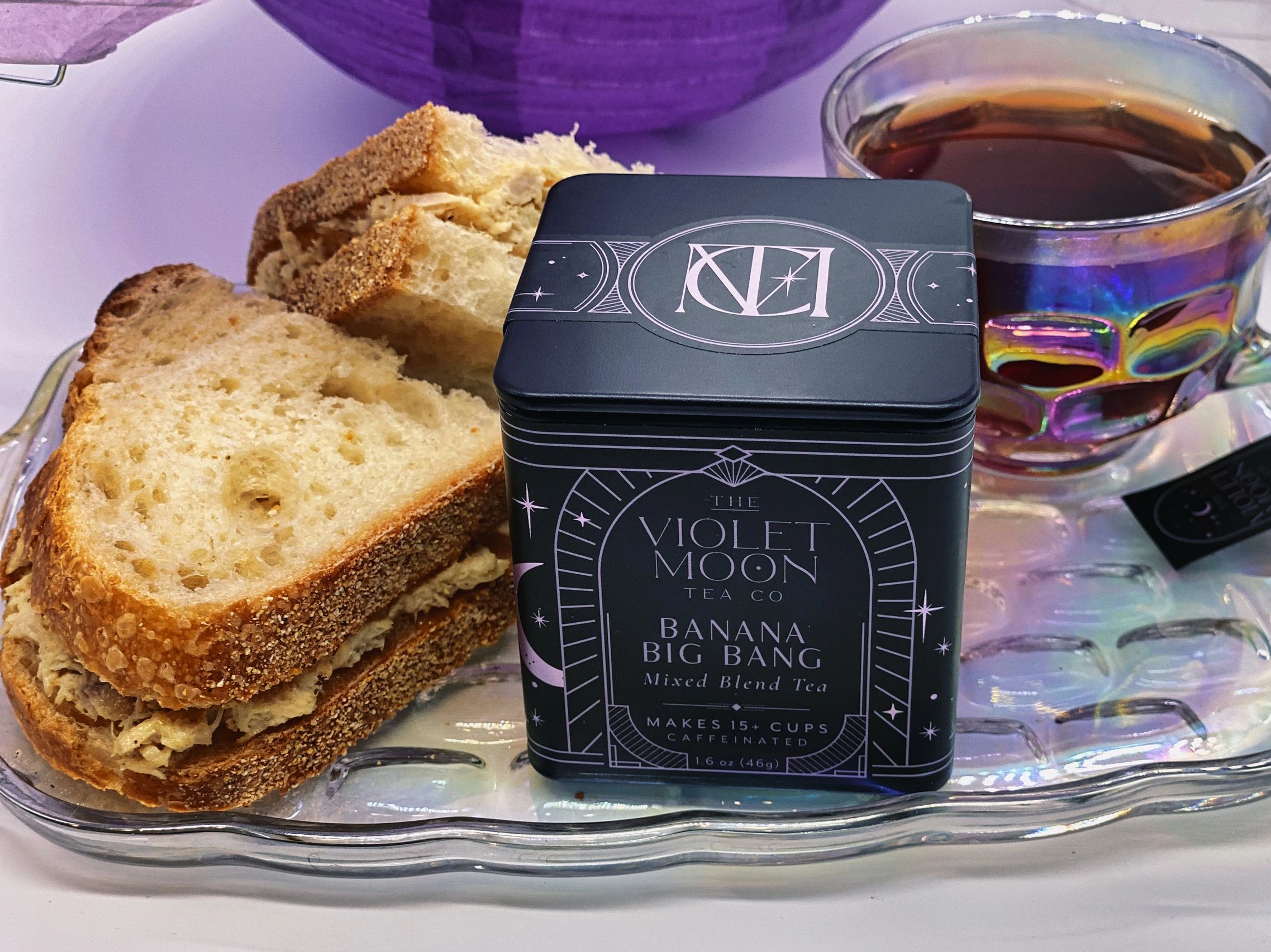 A black tin of Banana Big Bang tea on a plate with a cup of tea and a sandwich