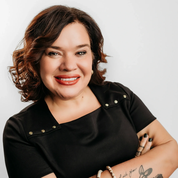 Woman with curly hair smiling, wearing a black shirt with tattoos and bracelets on her arm.