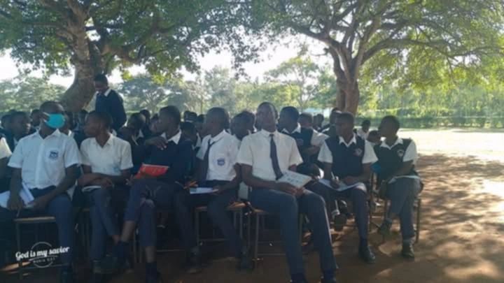 Students in uniform sitting outdoors under trees during a class or assembly.